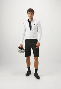 Hvit langermet sykkeltrøye med glidelås, kombinert med svarte sykkelshorts. Sykler med sko og en hvit hjelm holdt i hånden. Enkel, funksjonell design.