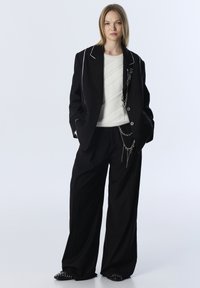 Zwarte oversized blazer met witte piping, gecombineerd met een gestructureerde witte sweater en brede zwarte broek met zilveren kettingaccenten.