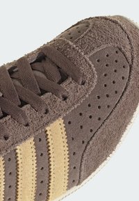 Brun mockasneaker med perforerad textur, med tre guldstripes. Bruna skosnören, texturerade paneler och vit gummisula.