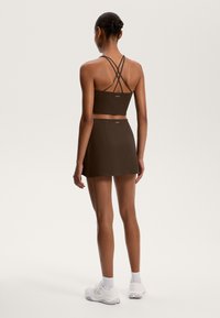 Rjav športni crop top z prečnimi naramnicami, v kombinaciji s skladiščenjem. Model nosi bele superge in gležnjarje. Smooth tekstura tkanine.