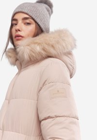 Jasnoróżowa kurtka puchowa z faux fur na krawędzi kaptura, z pikowanym wzorem, ściągaczami na mankietach i metką na rękawie z nazwą marki.