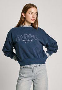 Mörkblå sweatshirt med "LONDON" tryck, ribbad mudd och fåll, bärs över en rutmönstrad skjorta med krage, kombinerad med ljusgrå jeans.