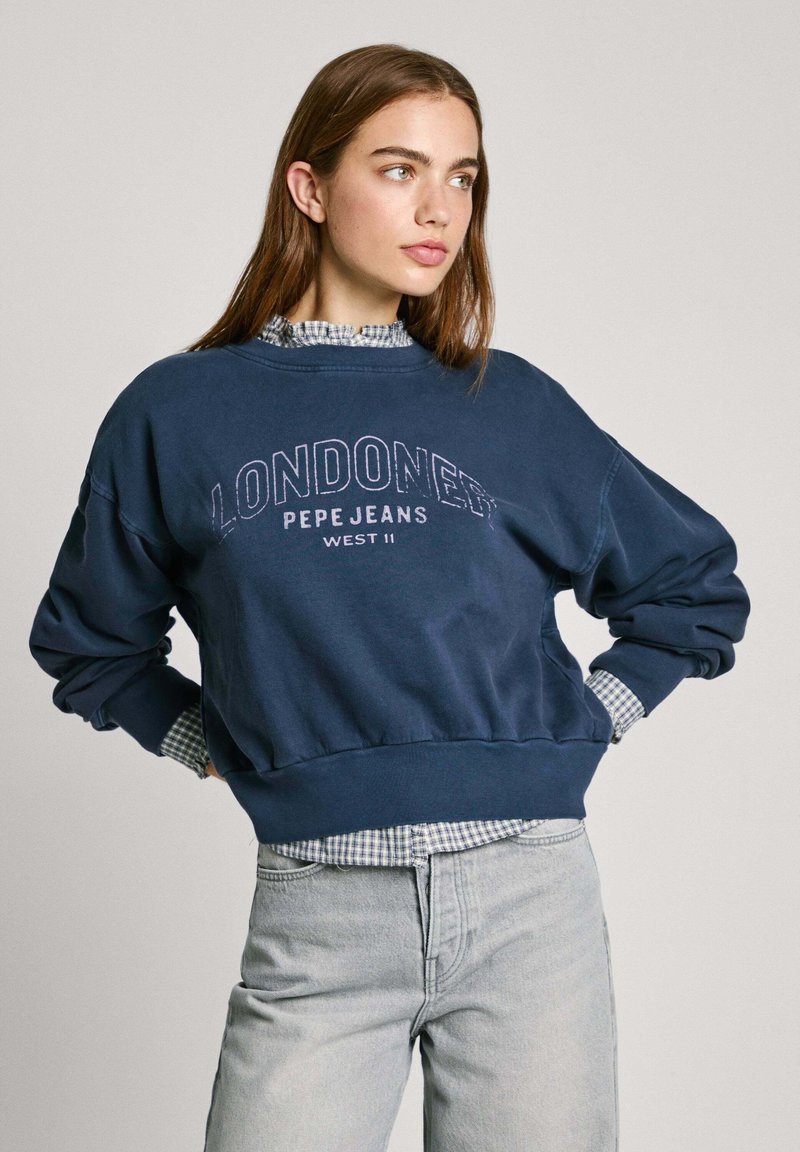 Mörkblå sweatshirt med "LONDON" tryck, ribbad mudd och fåll, bärs över en rutmönstrad skjorta med krage, kombinerad med ljusgrå jeans.