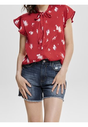 Nainen, jolla on päällään punainen kukallinen lyhythihainen pusero, jossa on rusetti kaulassa, ja siniset farkkushortsit, joista lahkeet ovat rispaantuneet. Hän seisoo kädet reisiä vasten lepäämässä.