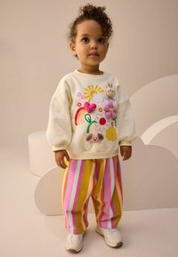 Sweatshirt blanc ample avec des appliqués colorés d'animaux et de fleurs. Pantalons de jogging rayés jaunes, roses et bleus. Baskets blanches.