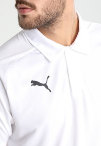 Polo bianco realizzato in tessuto traspirante con un logo Puma nero sul lato sinistro del petto. Design classico del colletto con patta.