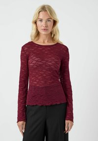 Top bordo a maniche lunghe realizzato in pizzo trasparente con motivi a onde testurizzati e bordi scallop, abbinato a pantaloni neri.
