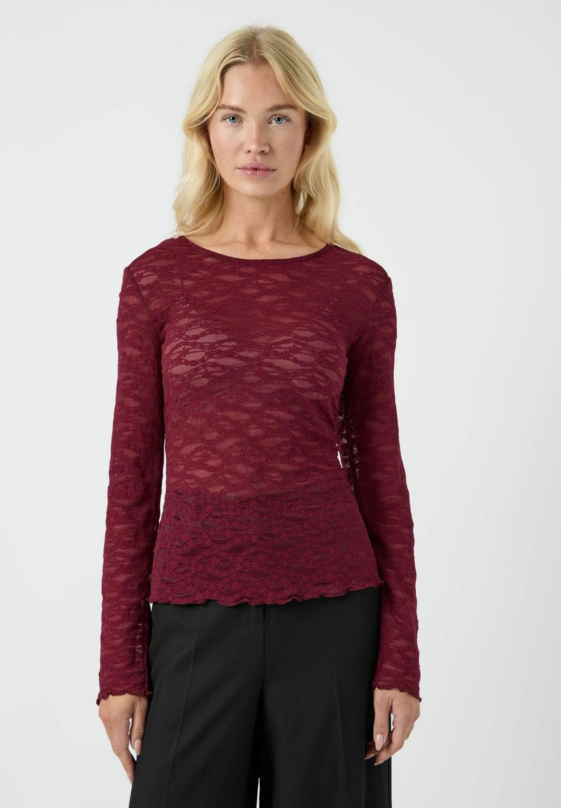 Top bordo a maniche lunghe realizzato in pizzo trasparente con motivi a onde testurizzati e bordi scallop, abbinato a pantaloni neri.