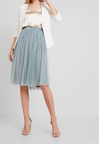Gonna di tulle azzurro chiaro con pieghe, abbinata a un top in satin dorato e a un blazer bianco. La modella indossa tacchi nudi con cinturini. Sfondo neutro.