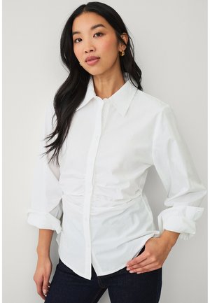 Młoda kobieta z długimi czarnymi włosami nosi białą koszulę zapinaną na guziki z podwiniętymi rękawami oraz ciemne dżinsy, pozując z jedną ręką na biodrze.