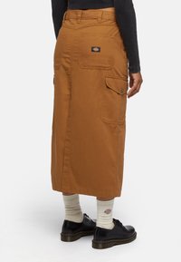Lunga gonna marrone realizzata in tessuto resistente, caratterizzata da due tasche cargo laterali, tasca posteriore con logo, indossata con scarpe nere e calze bianche.