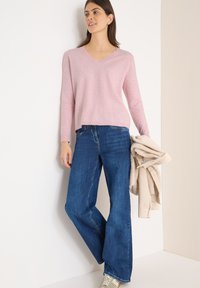 Suéter de punto en un suave tono rosa claro con escote en V y corte relajado, combinado con pantalones de mezclilla de pierna ancha en un azul oscuro. Ella sostiene un suéter de punto beige y lleva zapatillas grises en los pies.