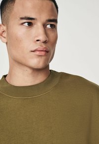 Sudadera verde oliva con un cuello redondo acanalado, que presenta una textura suave y un diseño simple; cortada de cerca para resaltar el escote.