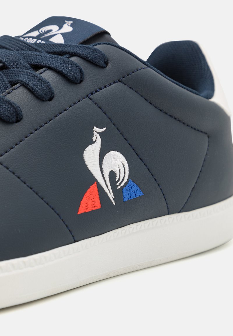 Zapatilla de piel sintética azul marino con suela de goma blanca, cordones oscuros y emblema de gallo bordado con detalles en rojo y azul.