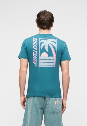 Bărbat purtând un tricou verde-albastru cu grafică roz cu palmier, apus și valuri și textul „Santa Cruz” pe spate, asortat cu pantaloni scurți de denim deschis la culoare.