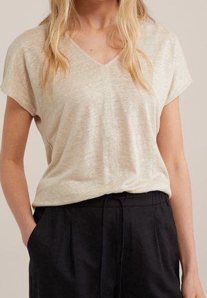 Frau trägt ein beigefarbenes Kurzarm-Shirt mit V-Ausschnitt, in schwarze Hosen mit Kordelzug gesteckt, Hände in den Taschen, steht vor neutralem Hintergrund.