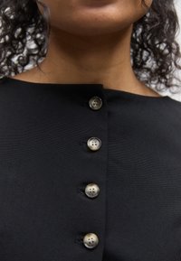 Close-up of a person wearing a black shirt with four pearlescent buttons down the front and curly dark hair visible.