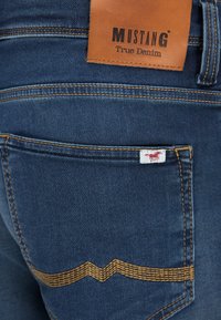 Tamsiai mėlyni džinsai su odine etiketė „MUSTANG True Denim“. Yra auksinė siuvimo detalė ir mažas raudonas logotipo ženkliukas ant galinio kišenės.