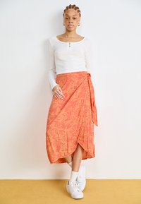 Oransje omslagsskjørt med blomsterprint, lett stoff, sideknute, kombinert med en hvit ribbestrikket topp med lange ermer og hvite sneakers.
