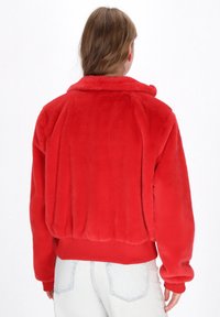 Jachetă roșie din blană artificială, cu guler, manșete și tiv ribbed, având o croială ușor scurtă. Prezintă o textură netedă și pufoasă.