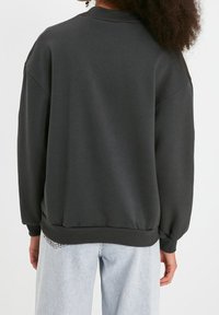 Sudadera gris oscuro con cuello redondo, puños y dobladillo acanalados; presenta un ajuste holgado y una textura de tela suave. Llevada con jeans de mezclilla claros.