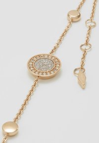 Pulsera de tono dorado con un centro circular de superficie texturizada, rodeado por un anillo de cristales transparentes, y un cierre con una etiqueta.