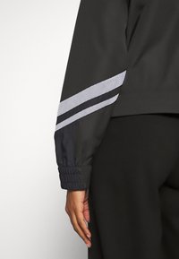 Musta urheilutakki, jossa on kontrastiväriset heijastavat raidat hihoissa, varustettu joustavilla hihansuilla ja sileällä, tekstuurisella kankaalla.