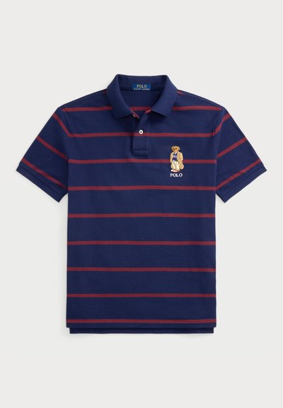 Marineblå polo med røde vandrette striber, korte ærmer og krave. Har et broderet bjørn-logo på venstre bryst.