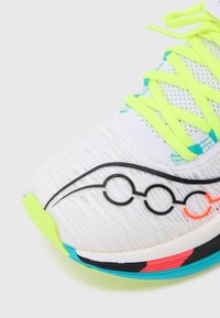 Zapatilla deportiva blanca con parte superior texturizada, cordones amarillos neón, detalles de líneas gráficas en negro y acentos en la suela de colores azul, rosa y verde.