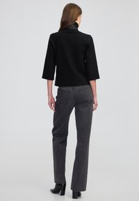 Vrouw die met de rug naar de camera staat, gekleed in een zwarte cropped jacket, donkergrijze rechte jeans en zwarte met hakken enkellaarsjes, tegen een effen achtergrond.