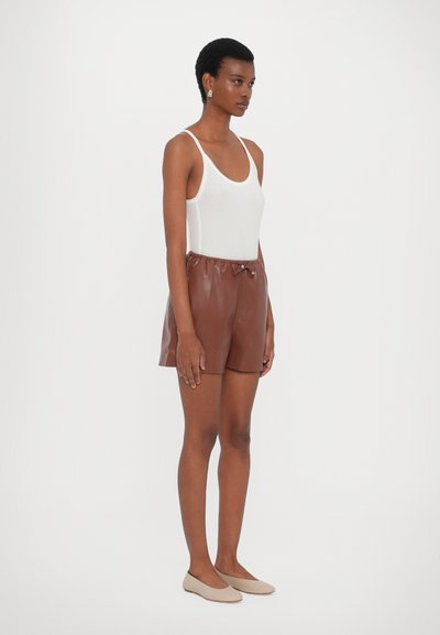Brune faux læder shorts med elastisk talje, der har en let glans. Bæres med en hvid tanktop og beige flade sko. Modellen står sidelæns.