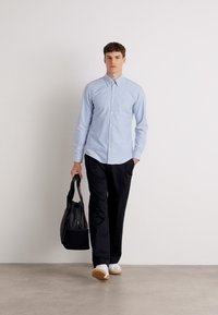 Šviesiai mėlynas marškiniai su užsegimu, derinami su juodomis kelnėmis, pasižymintys moderniu dizainu. Modelis laiko juodą maišą ir avėja baltas batus.