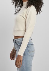 Sudadera crema con cuello alto, mangas largas ajustadas y dobladillo cropped, combinada con vaqueros azul claro. Textura de tela suave.