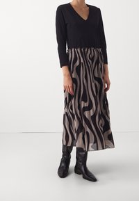 Svart V-ringad långärmad topp som kombineras med en midjelång plisserad kjol i beige och svart vågmönster, kompletterad med knähöga svarta stövlar.