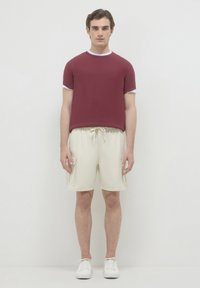 T-shirt bordeaux con rifiniture bianche, abbinato a pantaloni cargo beige con tasche laterali. Il modello indossa sneakers bianche, in piedi contro uno sfondo chiaro.