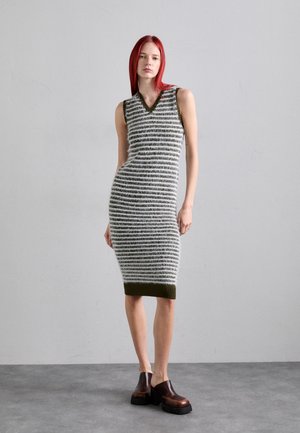 Robe sans manches en tricot avec des rayures horizontales grises et blanches, décolleté en V profond, coupe ajustée et bord contrastant en vert olive au niveau de l'ourlet. Chaussures brunes épaisses.