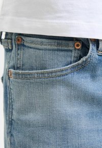 Jeans de mezclilla azul claro con un bolsillo frontal, detalles en botones de cobre y una textura suave. Dobladillo parcialmente cubierto por una camisa blanca.