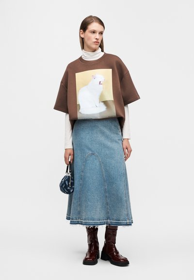 Bruine oversized trui met een afbeelding van een witte kat, gecombineerd met een lange blauwe denim rok en glanzende bruine laarzen tot aan de knie.