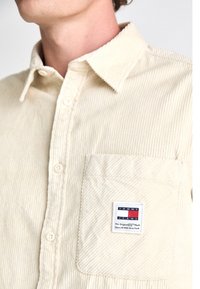 Camisa de pana beige con cuello abotonado, bolsillo frontal y una etiqueta de marca con rayas horizontales en azul, rojo y blanco.