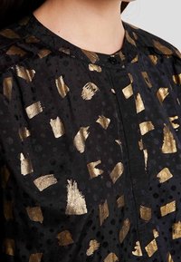 Blusa in tessuto nero con motivi astratti a pennellate metalliche oro, dotata di una piccola chiusura a bottone e polka dots testurizzati.
