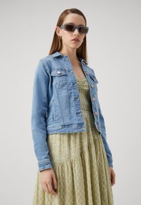 Chaqueta de mezclilla azul clara con botones de cobre, diseño ajustado y dos bolsillos en el pecho, combinada con un vestido verde con estampado.