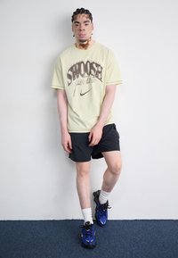 Svijetložuta oversized majica s tamnim grafičkim uzorkom i Nike logom, u kombinaciji s crnim šortsevima i plavim atletskim cipelama s crnim potplatima.