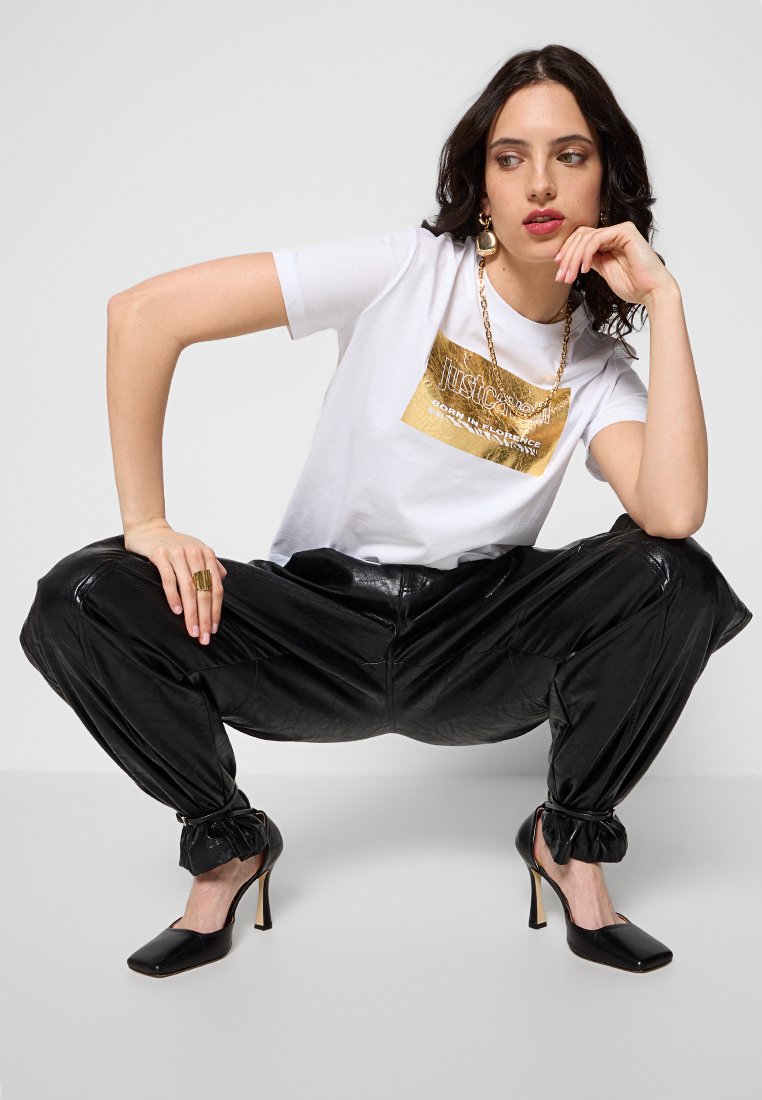 Camiseta blanca con un gráfico impreso en dorado, combinada con pantalones plisados negros y brillantes, y tacones de punta negra. Detalle de abertura en los puños de los pantalones.