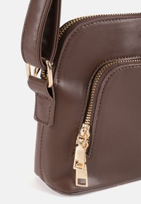 Esquina de bolso de mano de cuero marrón con cremalleras doradas y detalle de hebilla de correa ajustable sobre fondo blanco.