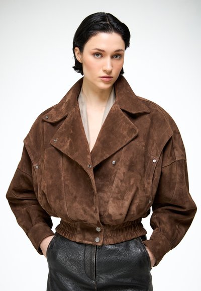 Personne aux cheveux courts et foncés portant une veste surdimensionnée en daim marron avec boutons-pression et un pantalon noir texturé, les mains dans les poches.