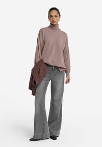 Kobieta w fioletowym swetrze z golfem, trzymająca bordową kurtkę, w połączeniu z szerokimi szarymi dżinsami i czarnymi, spiczastymi butami.