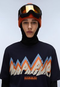 Tricou navy cu un grafic vibrant de munte în nuanțe de portocaliu, albastru și crem. Modelul poartă o căciulă tricotată portocalie și ochelari de schi. Este vizibil un fular negru.