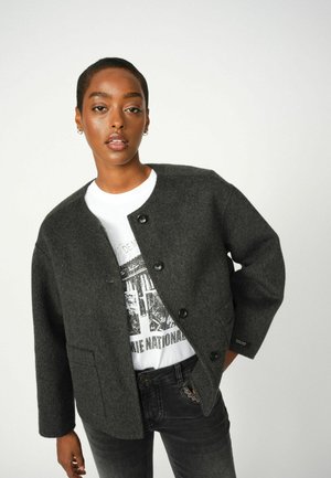Dark gray wool jacket with a round neckline, front buttons, and patch pockets, worn over a white graphic t-shirt and black jeans.