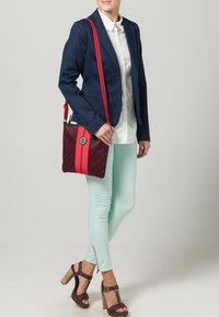 Červeno-modrá taška přes rameno s texturovaným vzorem, vybavená stříbrným logem a výraznými červenými pruhy. Modelka má na sobě tmavě modrý blejzr a mintové kalhoty.