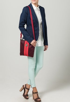 Červeno-modrá taška přes rameno s texturovaným vzorem, vybavená stříbrným logem a výraznými červenými pruhy. Modelka má na sobě tmavě modrý blejzr a mintové kalhoty.